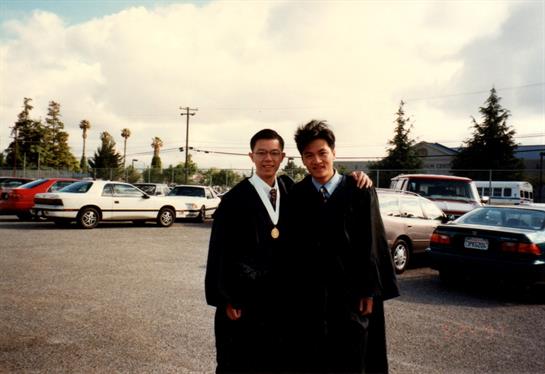 Two friends proudly pose together in graduation gowns in a parking lot under partly cloudy skies.