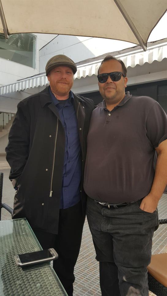 Two men stand side by side, smiling, in a cafe with modern architecture and bright sun.