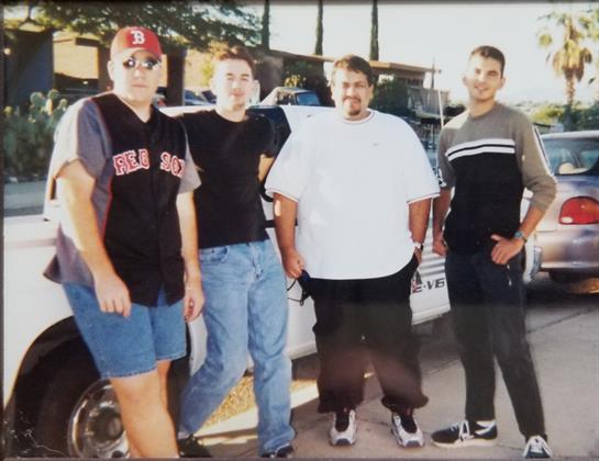 Four friends stand together smiling beside a parked car in a sunny location.