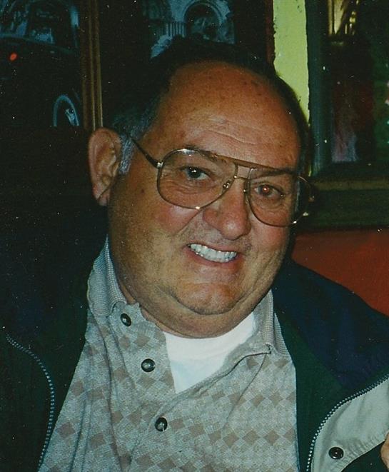 A cheerful man with glasses smiles warmly while seated indoors during a lively gathering.