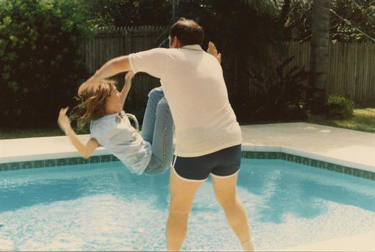 A child is being playfully lifted by an adult near a backyard pool filled with splashing water.