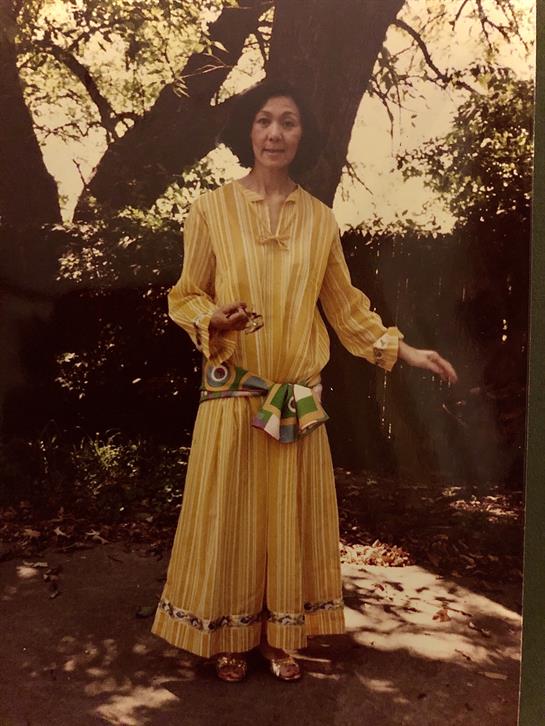 A woman stands gracefully in a flowing yellow dress, enjoying the sunny outdoor surroundings.
