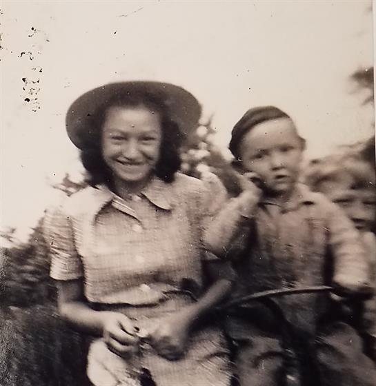 A joyful woman sits beside a young boy on a farm, both smiling and carefree, surrounded by greenery.