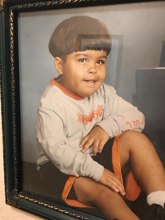 A young boy with a bowl haircut smiles while sitting on a chair, wearing a casual outfit.