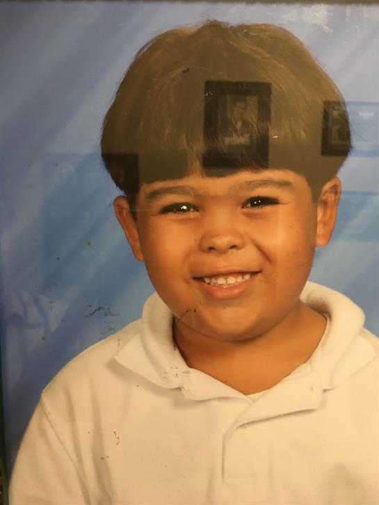 A boy with a unique haircut and white hoodie smiles brightly for a school portrait.