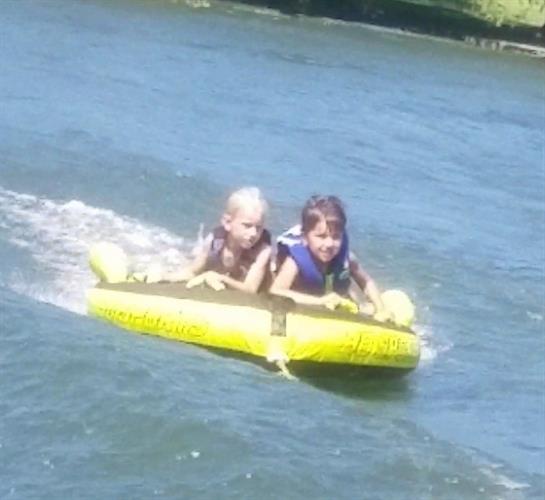 Two children happily ride a yellow inflatable boat on a sunny river during summer.