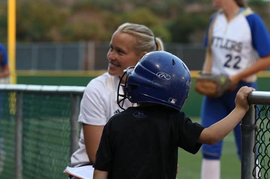 A coach smiles at a young player in a black shirt wearing a blue helmet during sports training.