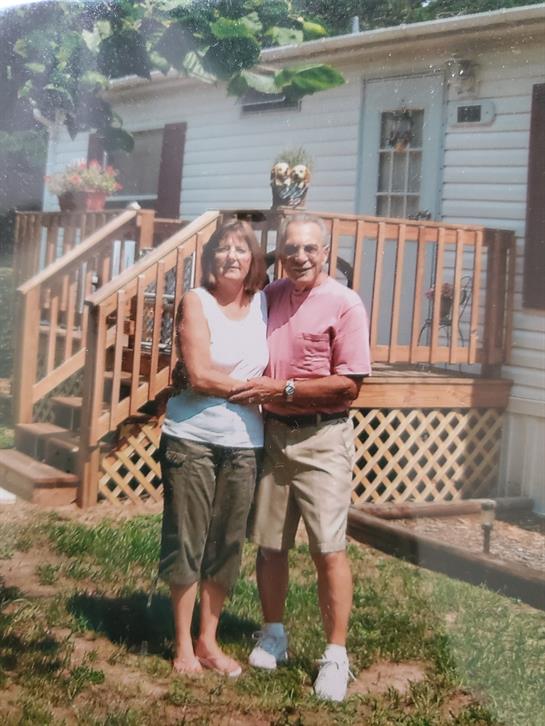 A senior couple stands happily together on the porch, framed by their charming home.