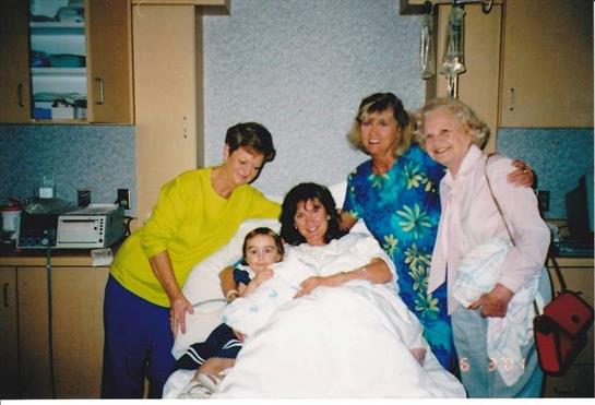 Five women and a child share joyful smiles in a hospital room, capturing a heartwarming moment.