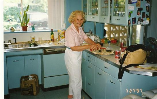 Older woman smiles while chopping vegetables in her retro-style kitchen filled with natural light.