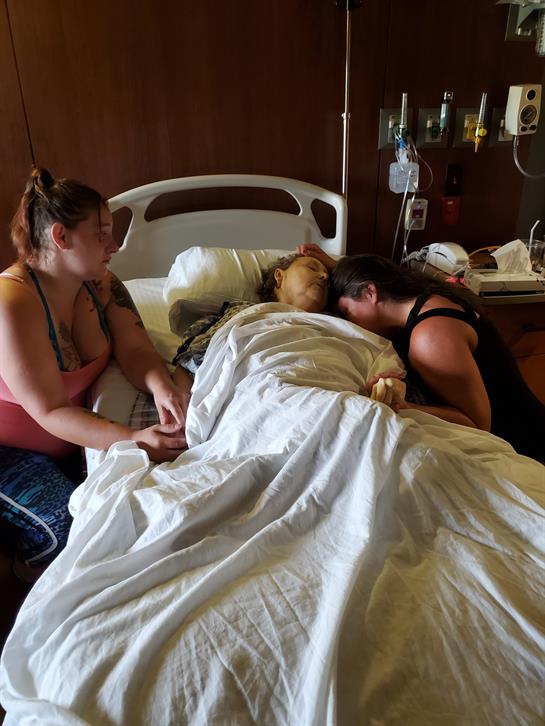Two women provide comfort to a loved one in bed at a hospital, sharing a heartfelt moment together.