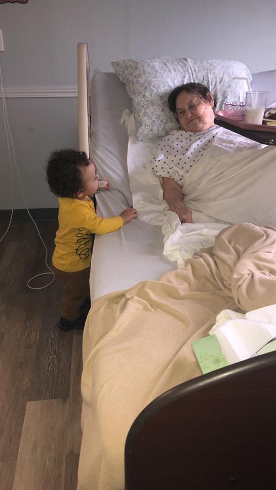 A young child stands by a hospital bed, interacting with their grandparent during a visit.