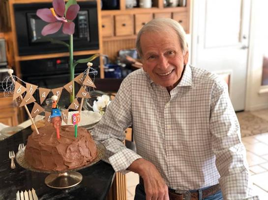 An elderly man smiles broadly next to a beautifully decorated cake in a warm kitchen setting.