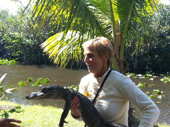 A person holding a crocodile near a riverbank surrounded by palm trees and vibrant foliage.