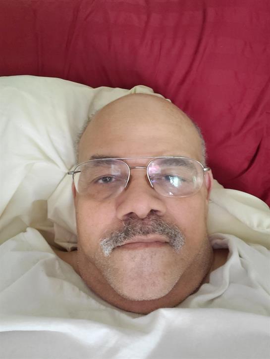 A man with glasses and a mustache is lying in bed, looking relaxed against a red backdrop.