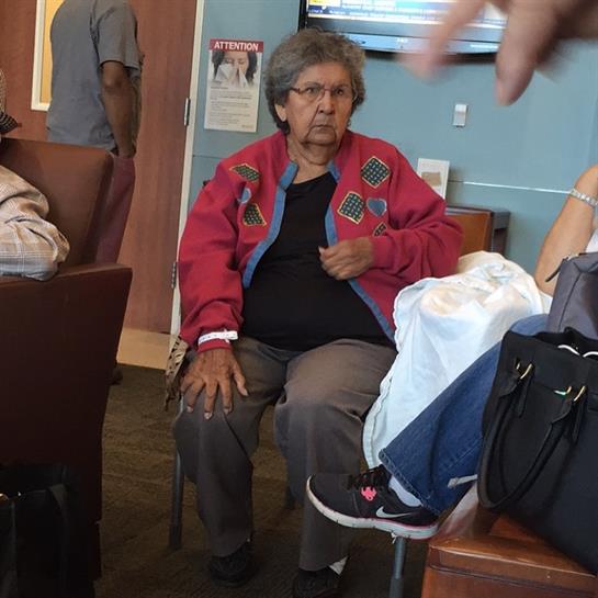 An elderly woman sits quietly in a waiting area, wearing a vibrant jacket while others chat nearby.