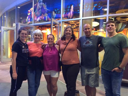 Six friends gather together, smiling, outside a vibrant restaurant after dinner during summer.