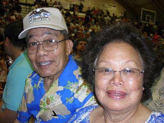 Two smiling seniors sit together in a busy community venue filled with people around them.
