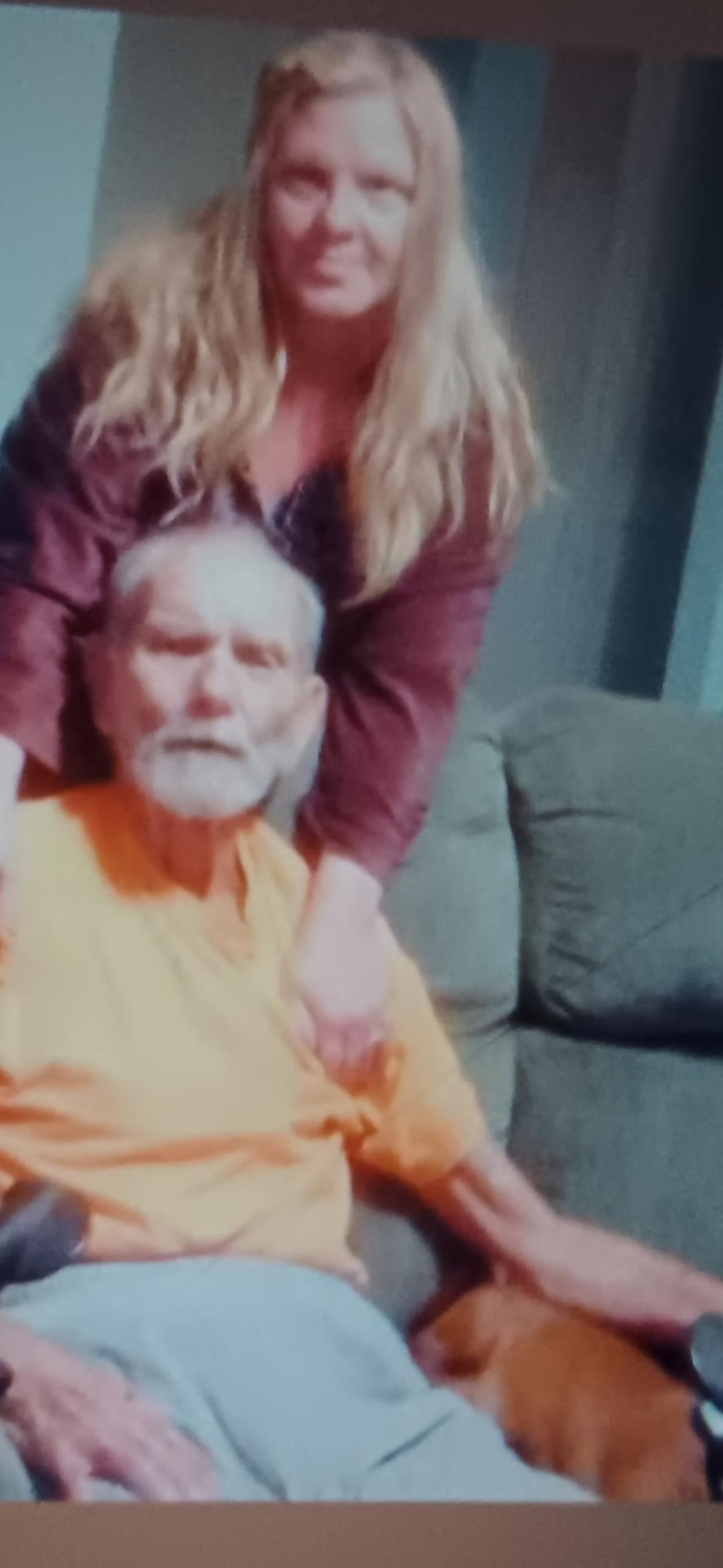 Elder man sits comfortably on a couch while a woman stands behind him, both sharing a joyful moment.