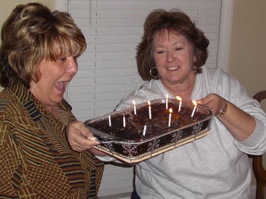 Two women joyfully surprise each other with a chocolate cake full of lit candles and laughter.