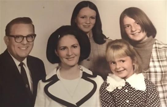 A group of five individuals poses together with warm smiles in a vintage 1970s setting.