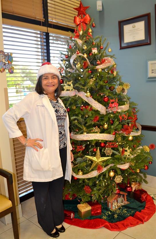 A healthcare worker stands joyfully next to a Christmas tree adorned with ornaments and lights.