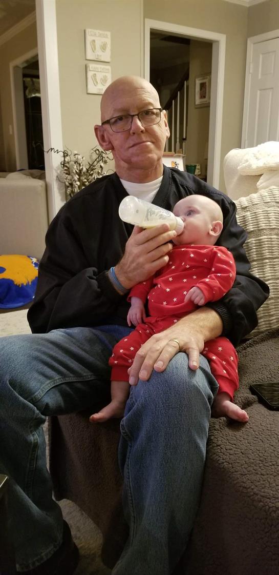 A grandparent sits on a couch while gently feeding a baby in a cozy living room.