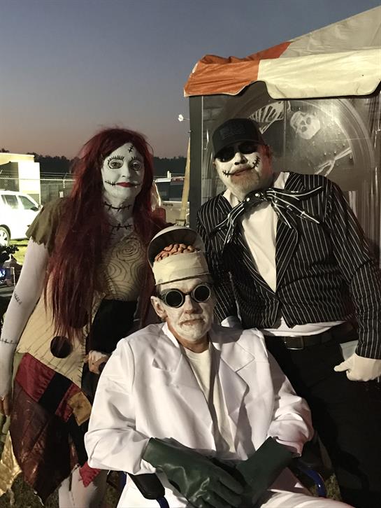 Three individuals in unique Halloween costumes pose together during an outdoor gathering at dusk.
