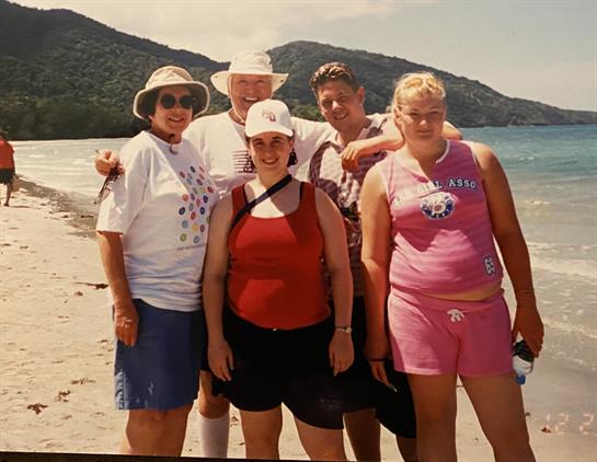 Group of five people smiling while standing on a sandy beach by the water during summer.