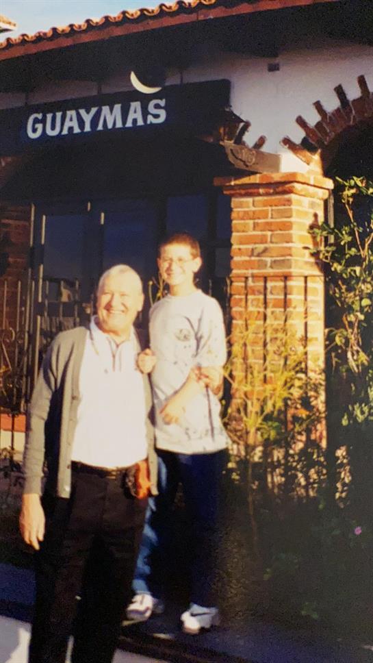 Elderly man stands beside young boy in a garden surrounded by plants during golden hour.