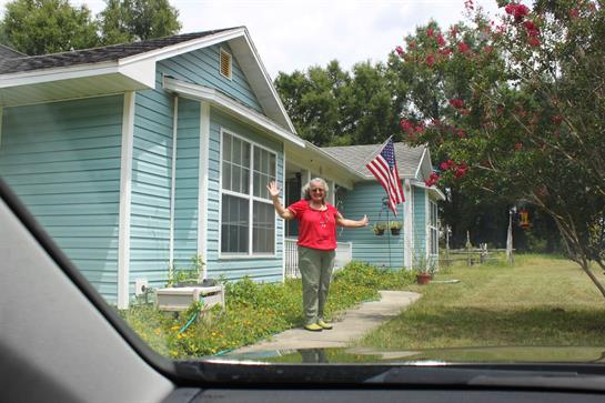 An elderly woman in a red shirt greets from her garden, surrounded by greenery and an American flag.