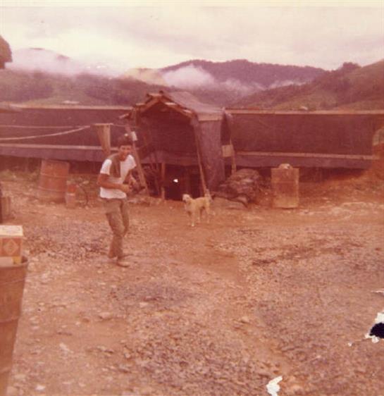 A man enjoys quality time with a dog near a simple shelter in a mountainous area at dusk.