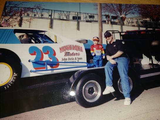 A man and a child in racing attire stand by a parked race car, enjoying a motorsport event together.