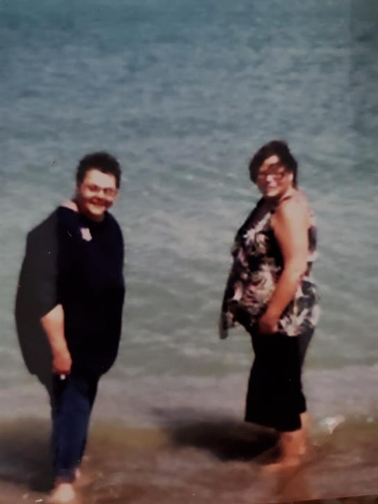 A couple stands by the water's edge, smiling and enjoying a sunny beach day with gentle waves.