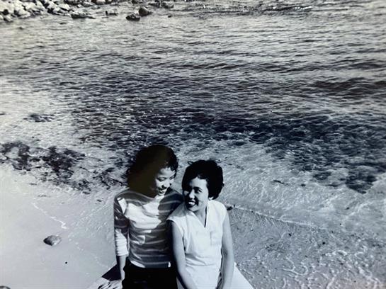 Two women sit close together on the sand, smiling and enjoying their time by the peaceful beach.