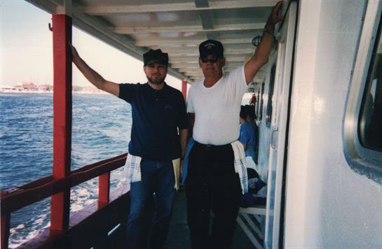 Two men pose casually on a boat deck with the ocean and sunlight in the background.