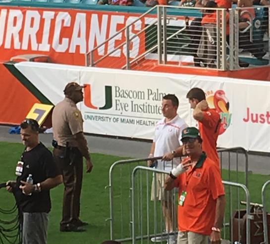 Fans in orange jerseys cheer as staff manage the crowd at a college football game in Miami.