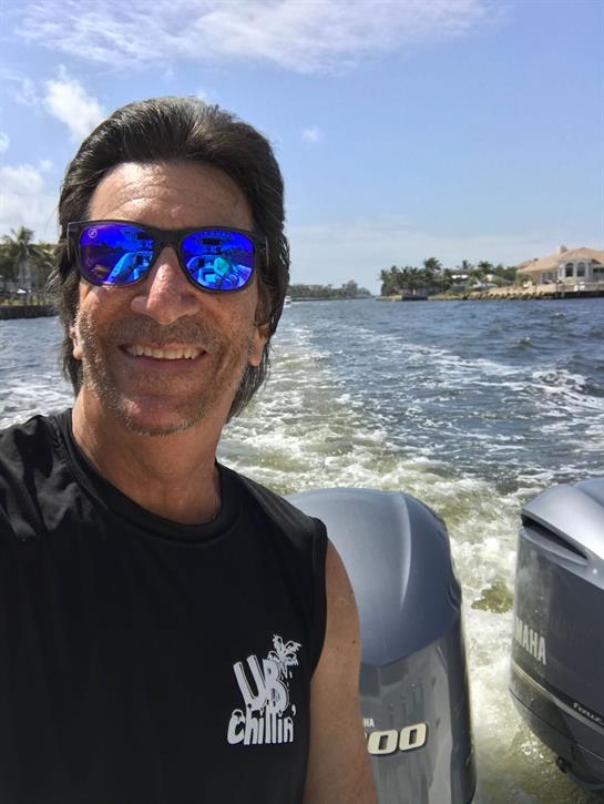 Man is smiling while cruising on a boat, surrounded by scenic waterfront homes and blue skies.