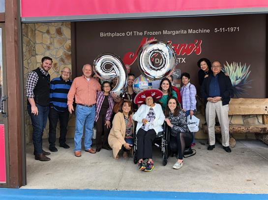 A group of family and friends joyfully celebrating a 90th birthday at a restaurant in Texas.