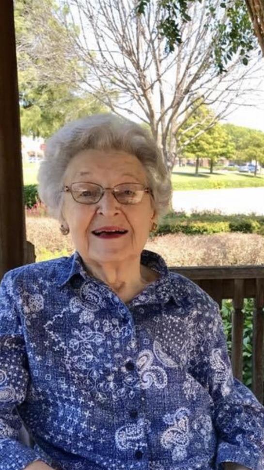 An elderly woman with gray hair enjoys a sunny afternoon in a park gazebo, smiling warmly.