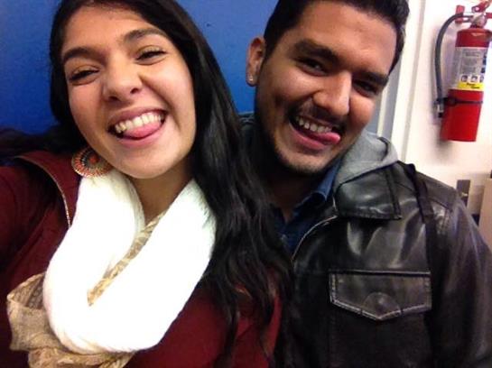 Two young adults share a fun moment, smiling and pulling faces while taking a selfie indoors.