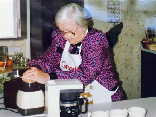 Senior woman brews coffee in a warm, inviting kitchen with floral wallpaper and simple decor.