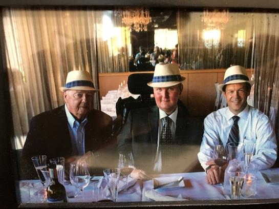 Three men, dressed in formal suits and straw hats, gather around a dining table, sharing a meal.