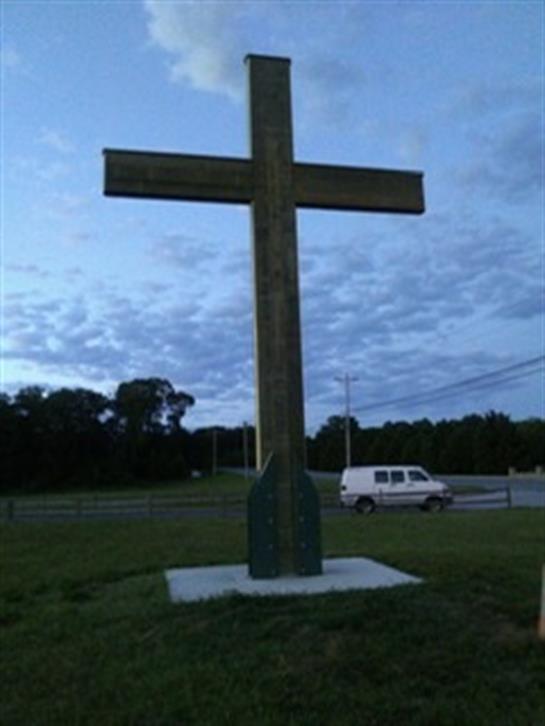 A tall wooden cross is situated in a grassy expanse adjacent to a road at dusk.