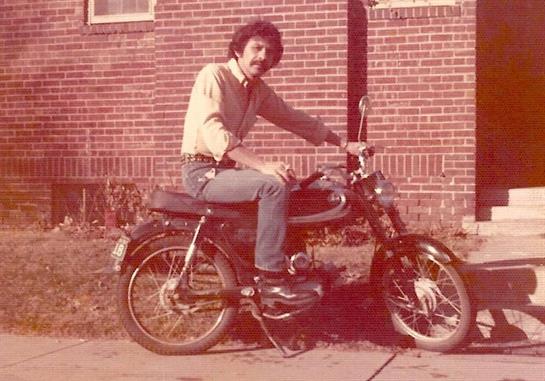 A young man poses confidently on a motorcycle, enjoying the sunny day in a vintage setting.