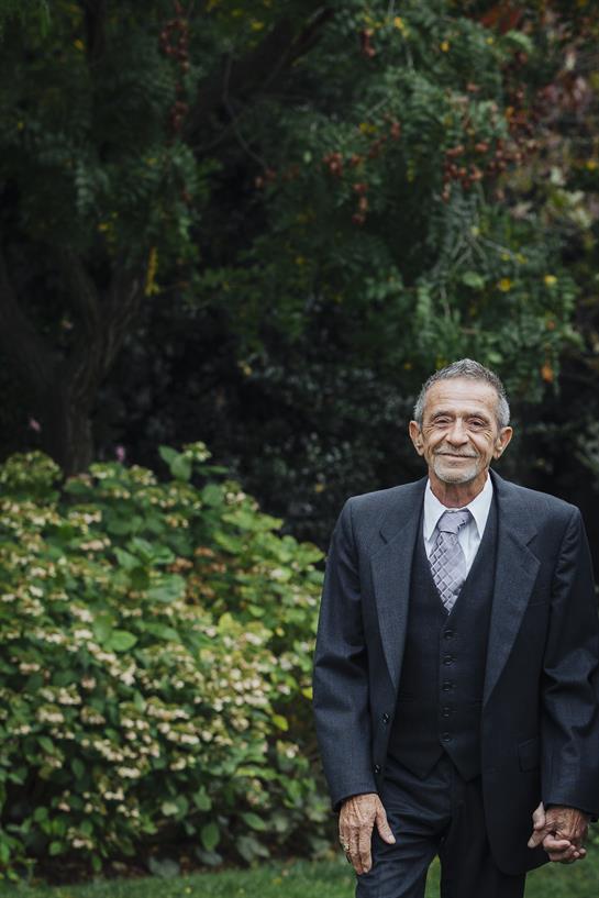 A cheerful older man in a formal suit stands confidently amidst vibrant greenery.