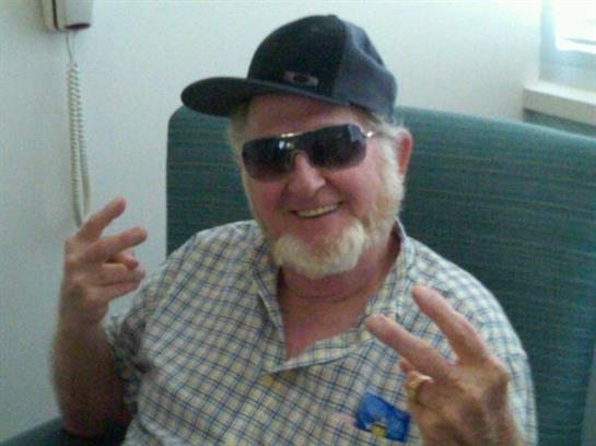 Smiling man sits casually on a chair, flashing peace signs and enjoying a relaxed moment indoors.