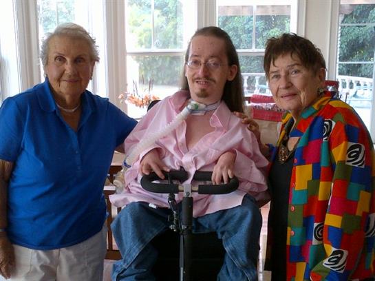 Two women smile while standing beside a man in a wheelchair at a cheerful indoor gathering.
