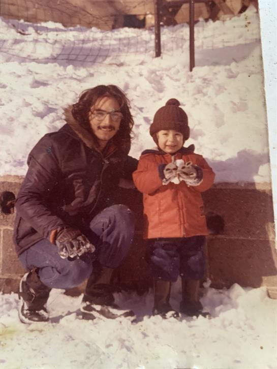 A father kneels next to his smiling child, both holding snowballs in a snowy yard.