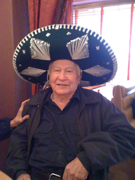 An elderly man smiles while wearing a large sombrero at a lively indoor celebration.